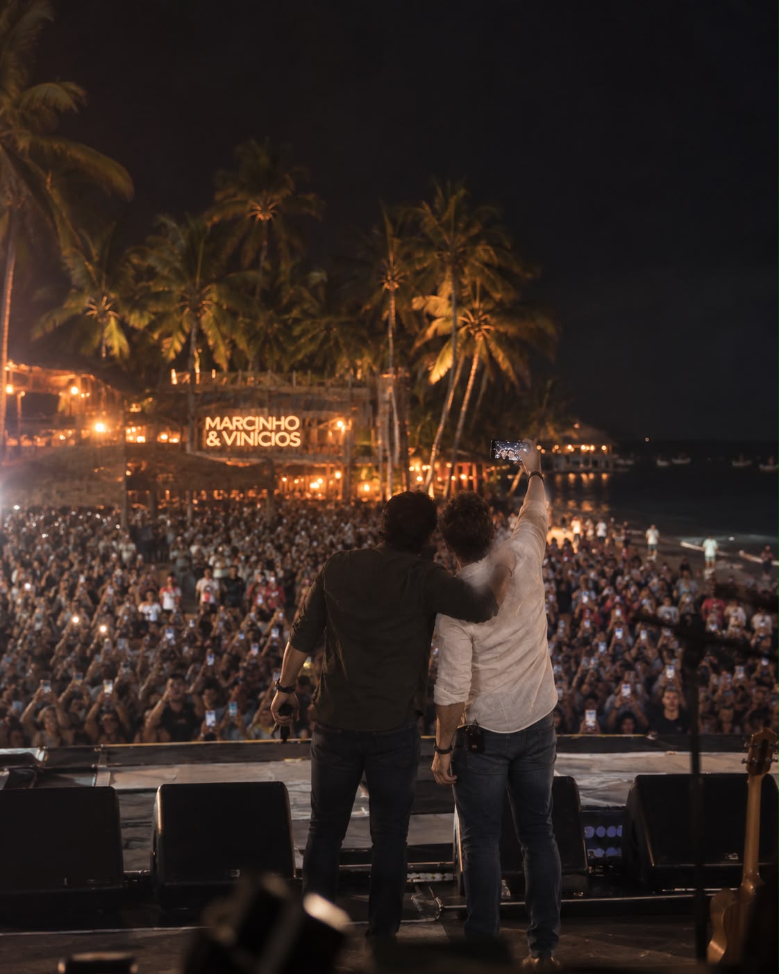 Marcinho e Vinícios de costas para o público, em show à beira-mar ao entardecer